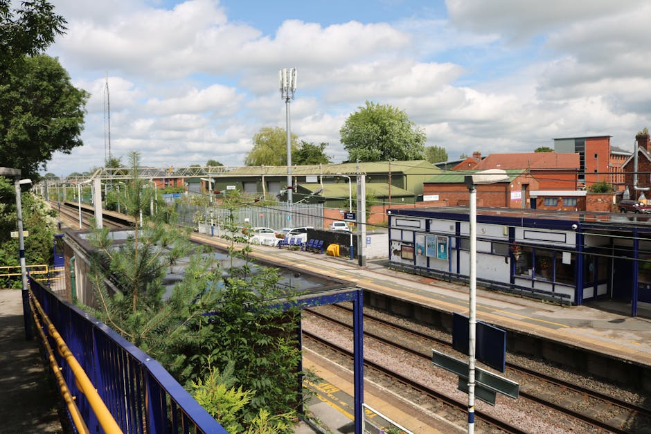 A view of Alperton Station with railway tracks running through the center of the image, bordered by a blue metal fence in the foreground. To the left, there is greenery including small trees and shrub