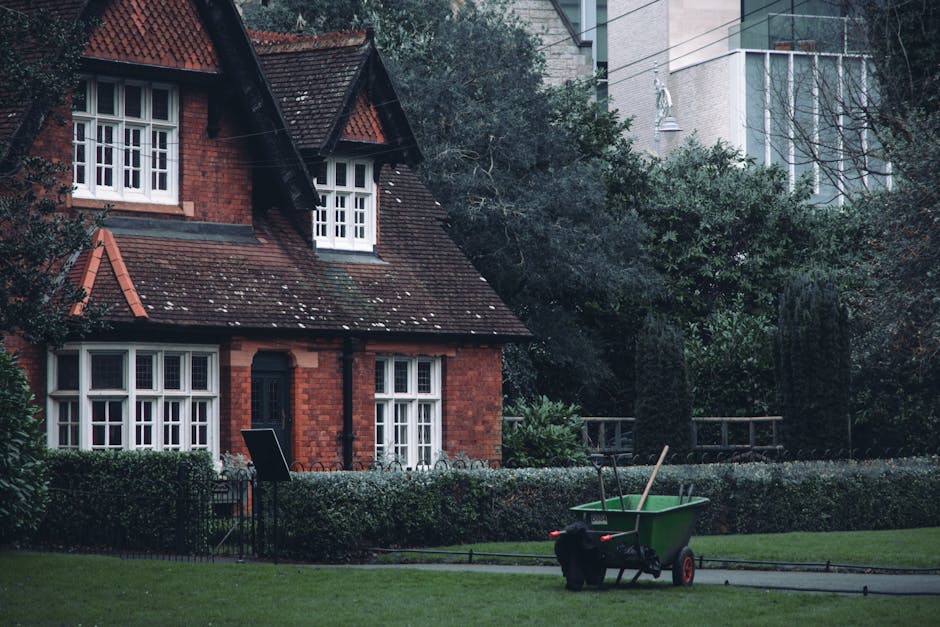 A residential garden scene featuring a well-maintained lawn with neatly trimmed grass, bordered by low hedges and mature trees with dense foliage. In the foreground, a green wheelbarrow filled with ga